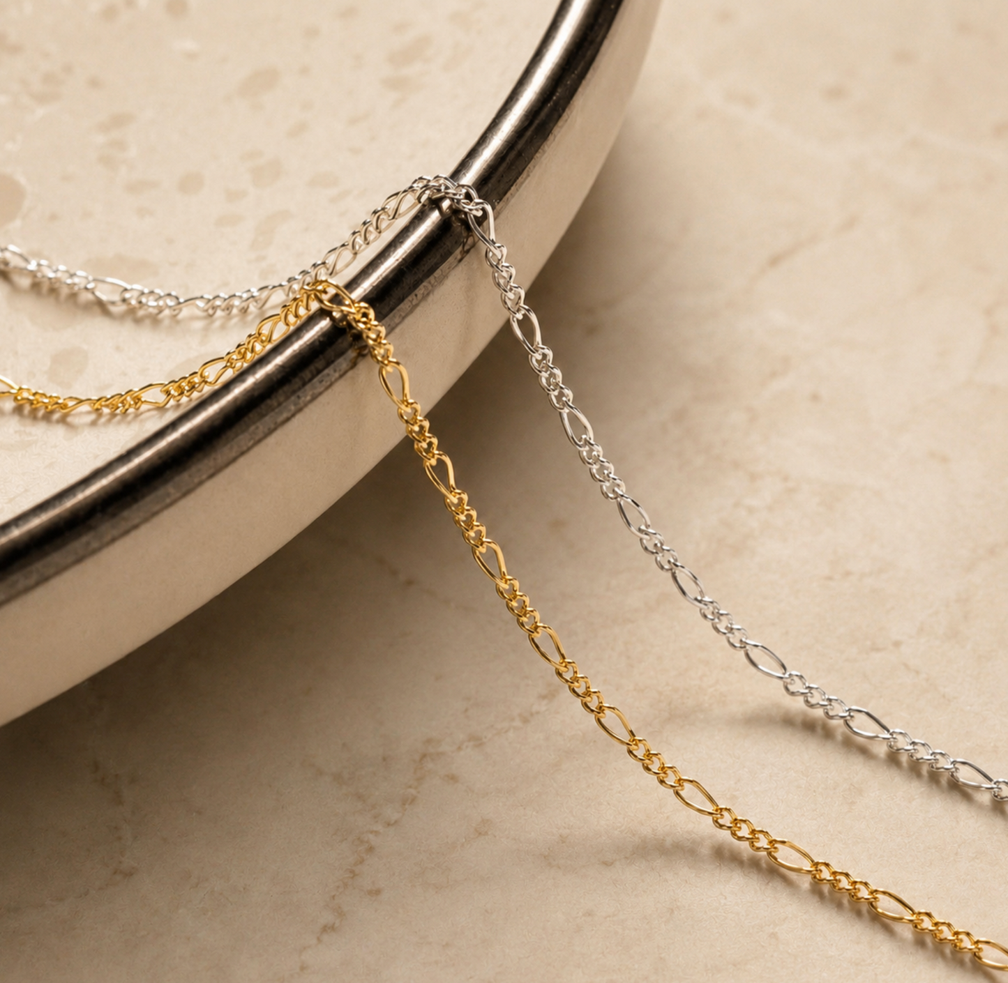 Two delicate metal chains, one gold and one silver, draped over the edge of a beige ceramic dish on a light stone surface.